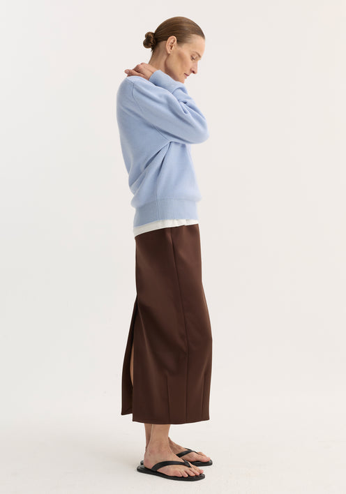 Model in baby blue compact knitted polo with collar and button placket, layered over white, paired with deep brown ankle-length skirt and black flat sandals.