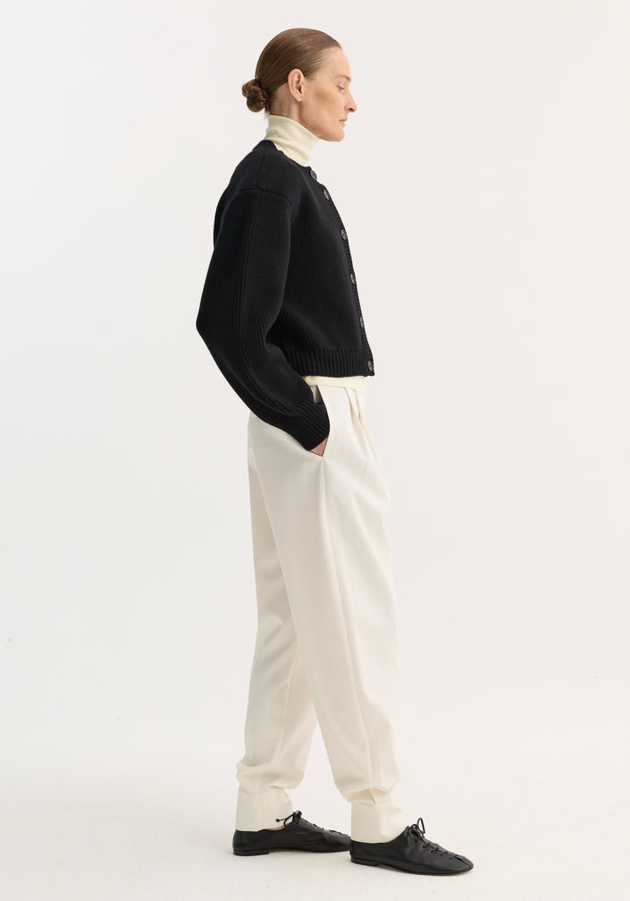 Model wearing Barrel leg trousers in off-white, featuring high-rise, relaxed tapered leg, double pleats, and ankle cuffs, styled with a black knit cardigan.