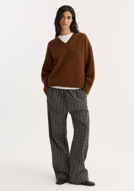 Model wears relaxed fit trousers in black/brown check with wide legs, elastic drawstring waistband, and side pockets. Paired with a V-neck sweater and white T-shirt.