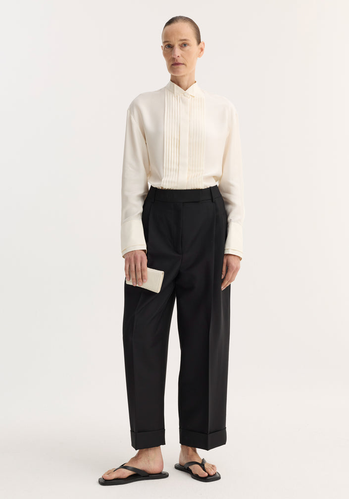 Model wearing high-waist turn-up trousers in black, featuring sharp pleating and wide legs, paired with an ivory blouse and minimal accessories against a plain backdrop.
