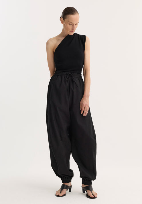 Person wearing black Balloon track pants with crinkled texture, elastic waistband, and cuffs, styled with a one-shoulder top and heeled sandals.