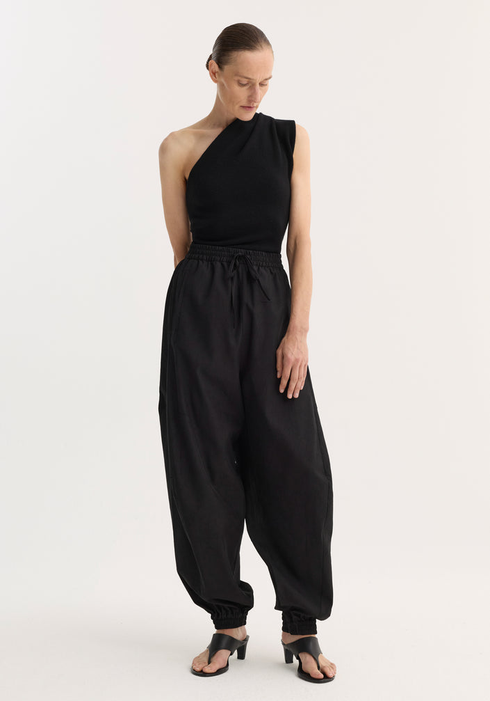 Person wearing black Balloon track pants with crinkled texture, elastic waistband, and cuffs, styled with a one-shoulder top and heeled sandals.