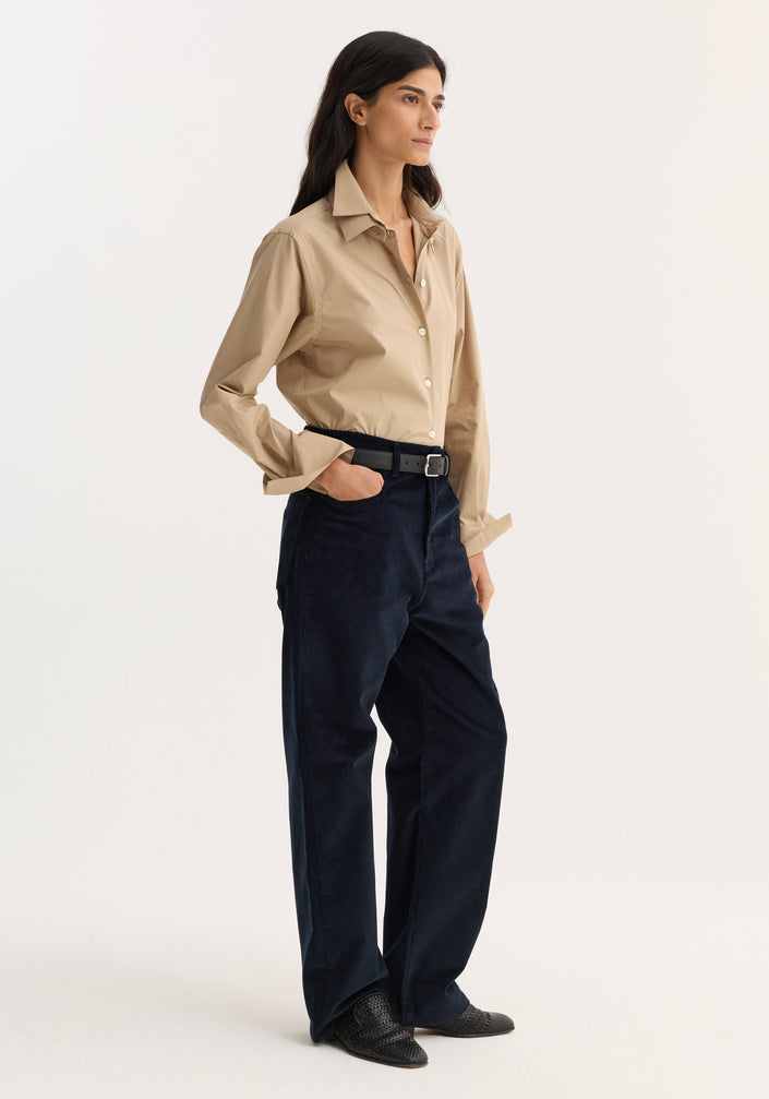 Woman wearing Relaxed corduroy trousers in navy, styled with a beige button-down shirt, black belt, and shoes, standing against a plain background.