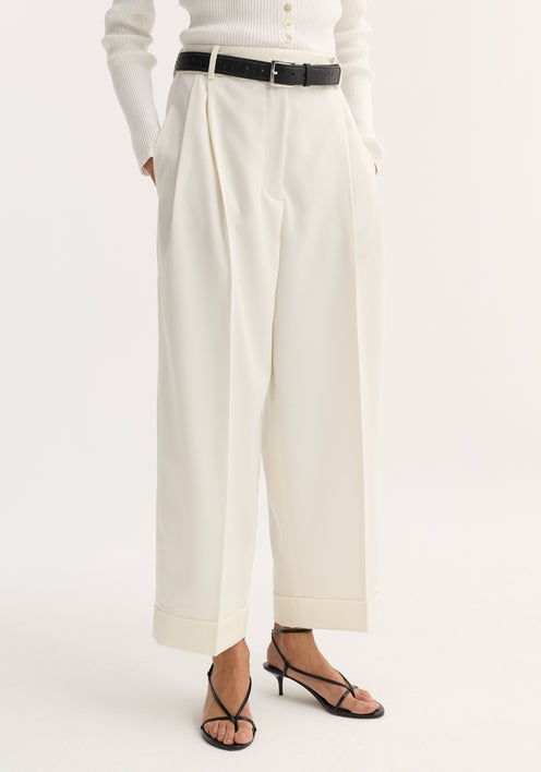 Woman wearing high-waist turn-up trousers in off-white with sharp pleats, paired with a ribbed polo-style top, black belt, and strappy sandals.