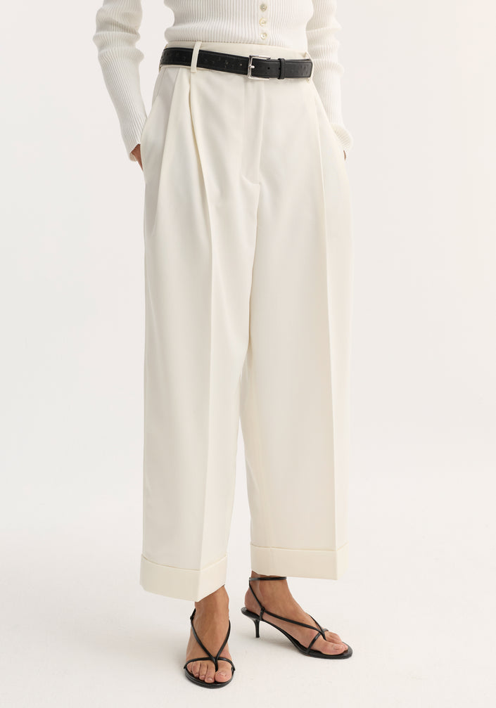 Woman wearing high-waist turn-up trousers in off-white with sharp pleats, paired with a ribbed polo-style top, black belt, and strappy sandals.