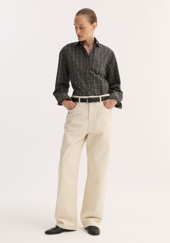 Person in dark plaid shirt with hands in pockets, showcasing Baggy fit denim | ecru with a relaxed, wide-leg, low-waist design and black slip-on shoes.