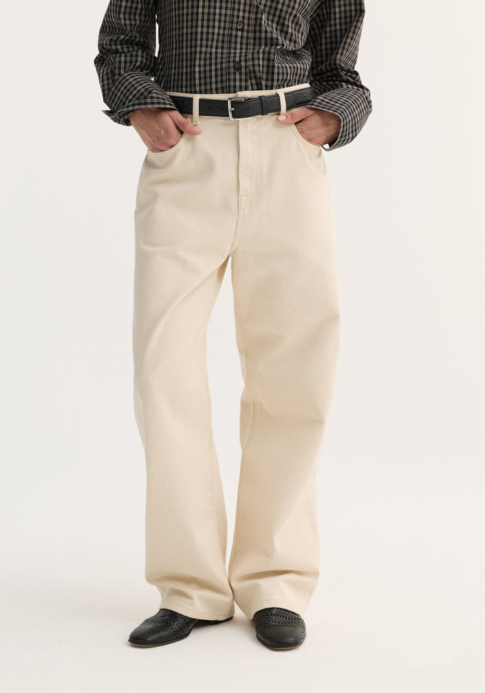Person in dark plaid shirt with hands in pockets, showcasing Baggy fit denim | ecru with a relaxed, wide-leg, low-waist design and black slip-on shoes.