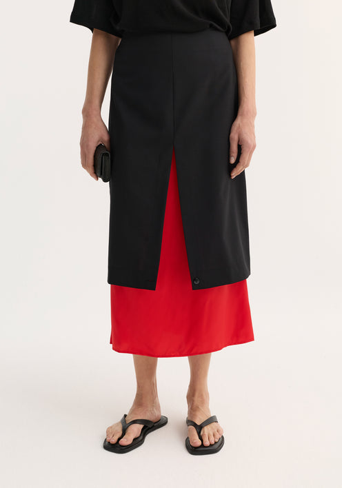 Model in a Contrast upside-down skirt | black featuring a black pencil skirt with red underlay, front slit, black top, and flip-flop sandals, holding a clutch.
