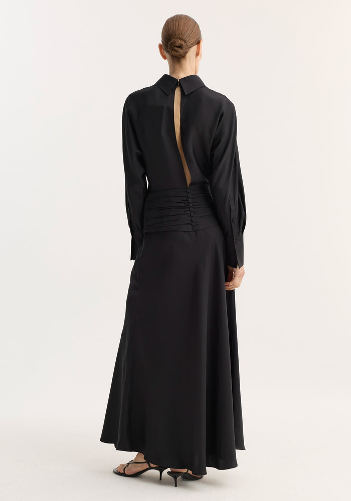 Model in Tuxedo silk dress | black, featuring a flowing design with a pleated waistband, reversed collar, and open back. Hair in bun, with strappy sandals.