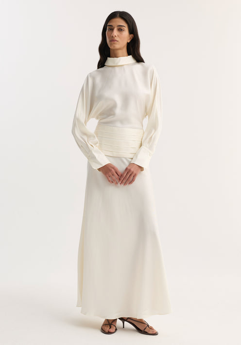 Model in Tuxedo silk dress | cream, featuring a high-neck blouse, pleated waistband, A-line skirt, and open back with cummerbund and side zip detailing.