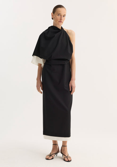 Model in a floor-length Draped open back dress | black, featuring an asymmetrical one-shoulder design, contrasting white lining, and strappy heeled sandals.