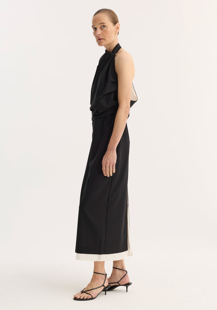 A slender model in a Draped open back dress | black, with a halter top, open back, side slit long skirt, and strappy sandals, posed in profile.