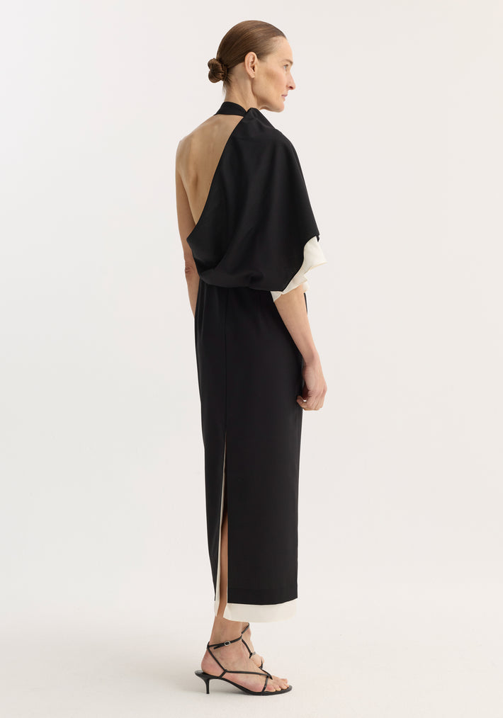 Model in profile wearing the Draped open back dress | black, featuring a sleeveless design, single draped shoulder, contrasting hem, and back slit, paired with black sandals.