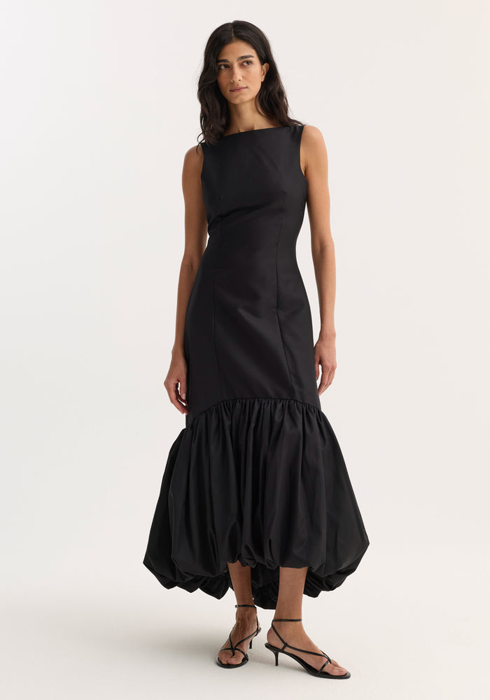 Model in a Voluminous dress | black, showcasing a sleeveless, form-fitting design with a boat neckline and a peplum hem, paired with strappy heeled sandals.