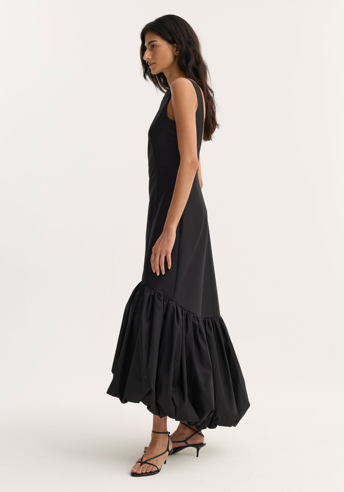 Voluminous dress | black: Slim woman in profile wearing a sleeveless, form-fitting black maxi dress with a boat neckline and sculptural peplum hem, paired with black heeled sandals.