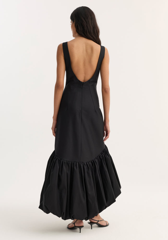 Voluminous dress | black: Woman in a sleeveless, scoop-back, form-fitting black dress with a peplum hem, standing against a neutral background in black strappy sandals.