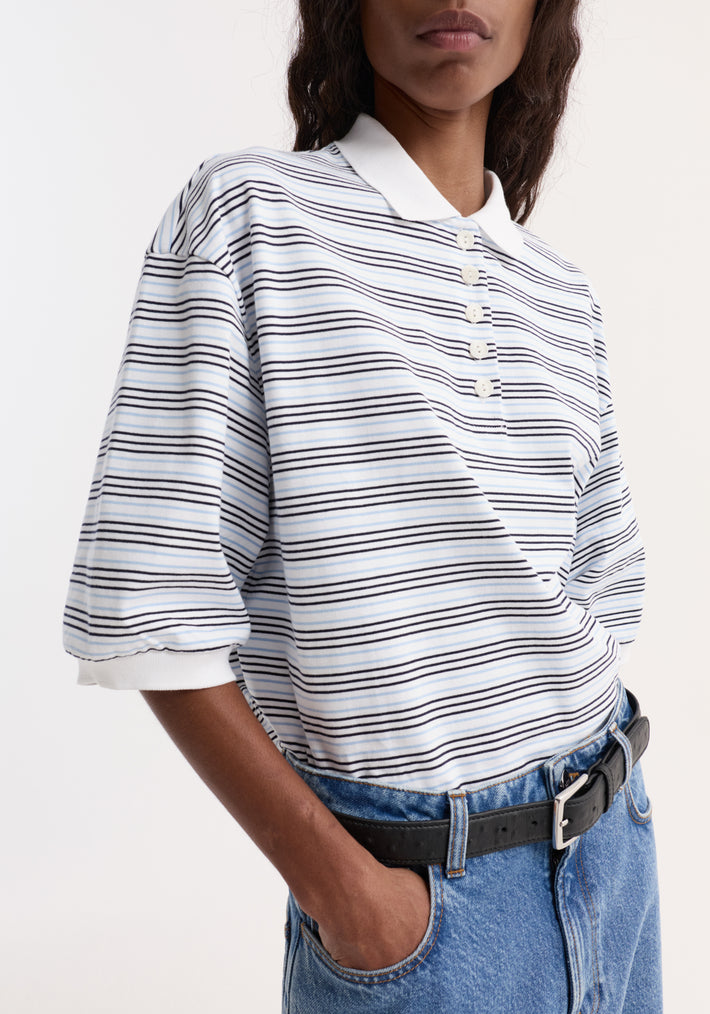 Striped oversized polo in white and blue multi-stripe, featuring a white collar, four-button placket, and relaxed fit, paired with mid-wash jeans and black belt.