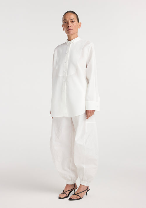 A model wearing a long-sleeve tuxedo shirt | optic white with a stand-up collar and concealed buttons, paired with loose white pants and black strappy heels.