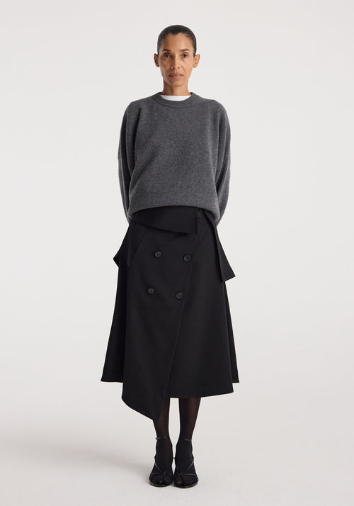 A woman wearing a black Deconstructed trench skirt | black with an asymmetrical double-breasted design paired with a dark gray oversized sweater and black shoes, standing against a plain white background.