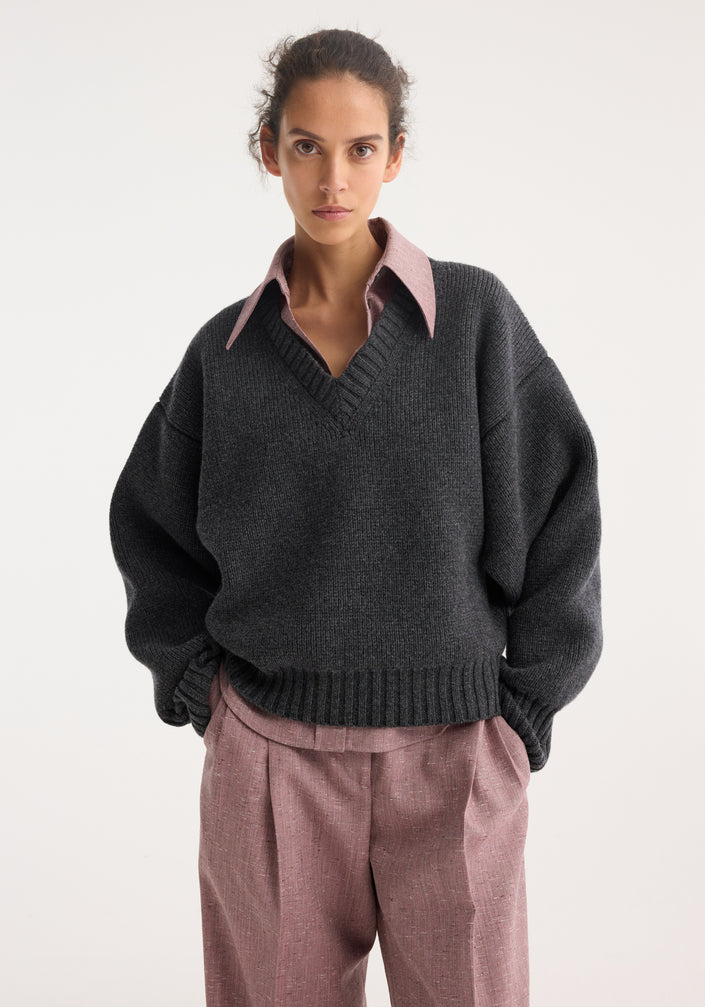 Model wearing Sculptural compact V-neck mid grey melange sweater over pink shirt with pointed collar, paired with pink wide-leg trousers and brown suede flats.