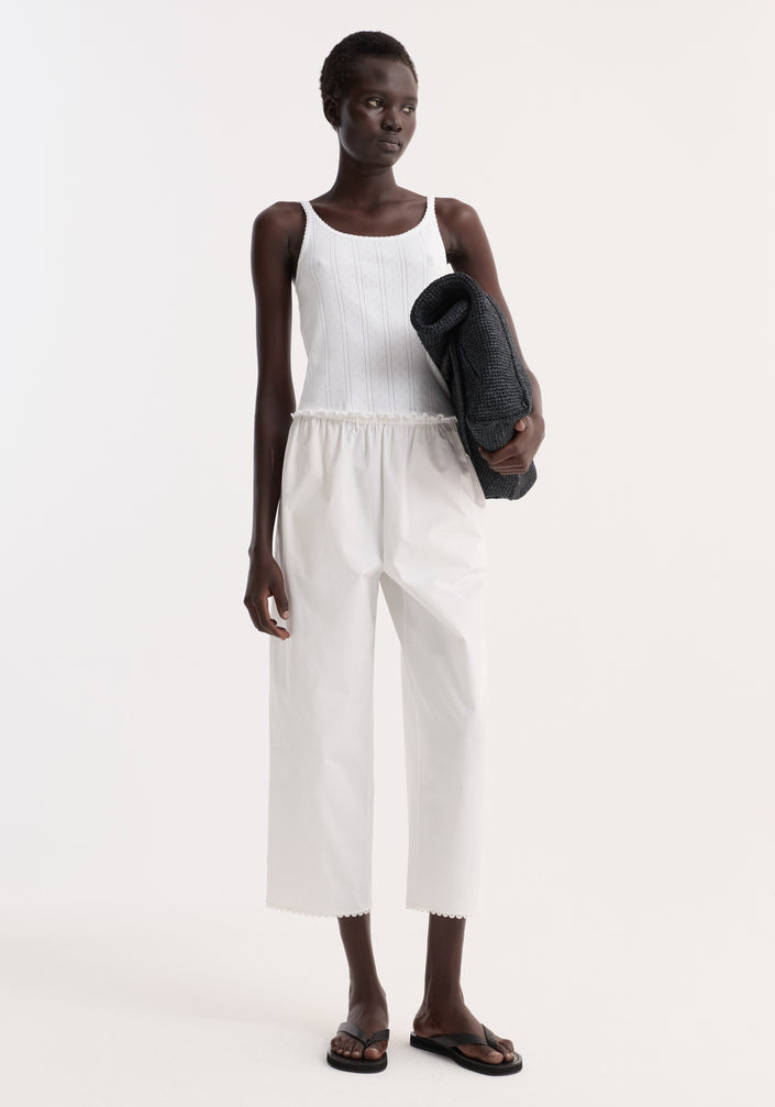 Model wearing Pointelle tank in off white, featuring a ribbed, sleeveless design with lettuce hem, paired with high-waisted trousers and holding a dark gray knit sweater.