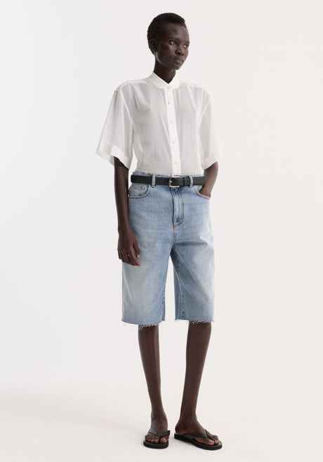 Model in an Embroidered muslin shirt | optic white, featuring sheer cotton, drop short sleeves, and embroidery, paired with light-wash denim shorts and a black belt.