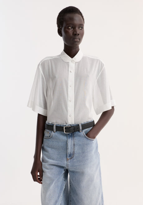 Model in an Embroidered muslin shirt | optic white, featuring sheer cotton, drop short sleeves, and embroidery, paired with light-wash denim shorts and a black belt.