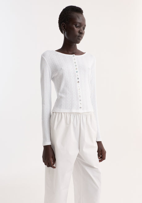 Model wearing the Pointelle cardigan in off white, showcasing its scalloped neckline, mother-of-pearl buttons, paired with high-waisted trousers and black sandals.