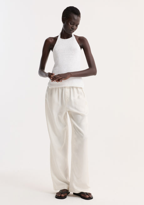 Model wearing Chunky knit halter top in optic white, featuring side slits and button closure, paired with loose trousers and black slide sandals.