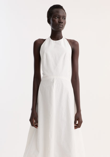 Woman wearing Halter textured cotton dress | off white, featuring a sleeveless halter-neck, low back, A-line skirt, paired with black strappy heeled sandals.