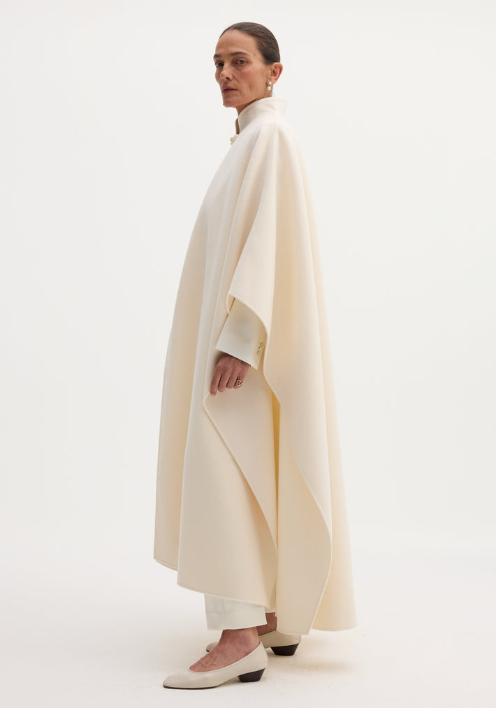 Model in oversized Pankou closure poncho, off-white, with stand collar, paired with white trousers and neutral pumps, showcasing refined layered style.