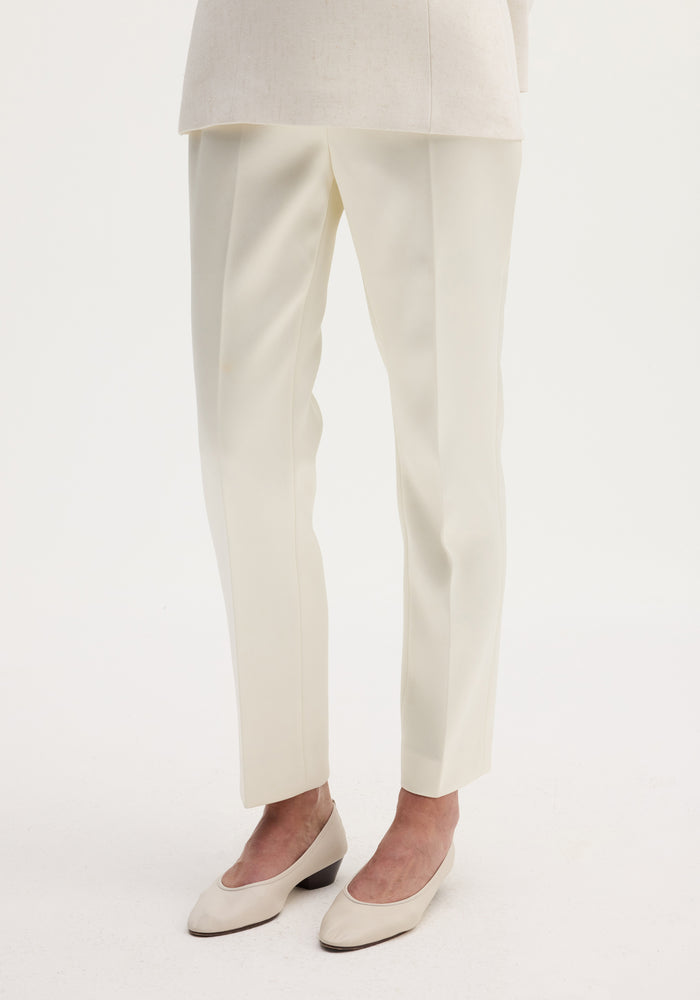 Cigarette leg satin trousers in ivory, featuring a high-rise fit and sharp center crease, paired with matching block-heeled pumps on a plain background.