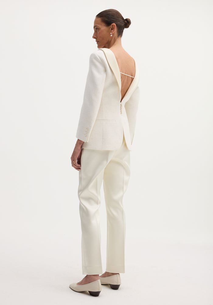 Woman in Reversed blazer | winter white, showcasing a deep cowl back with thin strap, satin lapel, welt pockets, button closures, and straight-leg trousers.
