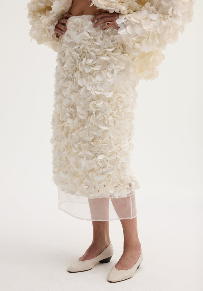 Handmade embellished skirt in ivory, featuring a voluminous knee-length design with delicate fabric appliqués and a refined silk organza hem, modeled with cream low-heeled pumps.