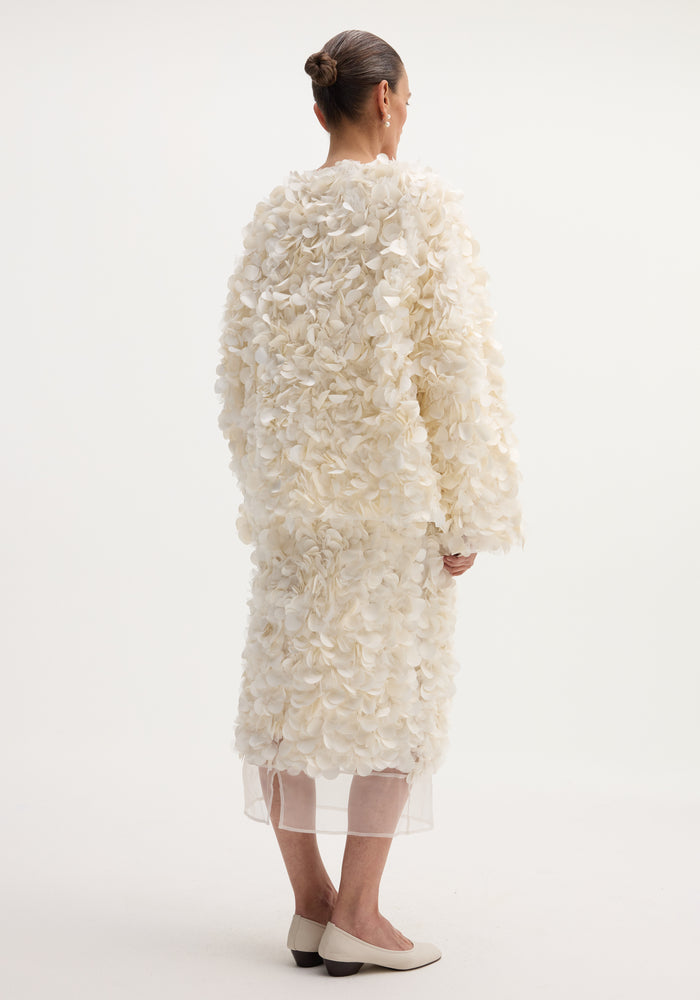 Back view of a woman wearing a Handmade embellished jacket in ivory, featuring petal-like appliqués, over a matching skirt, showcasing the jacket's voluminous design.