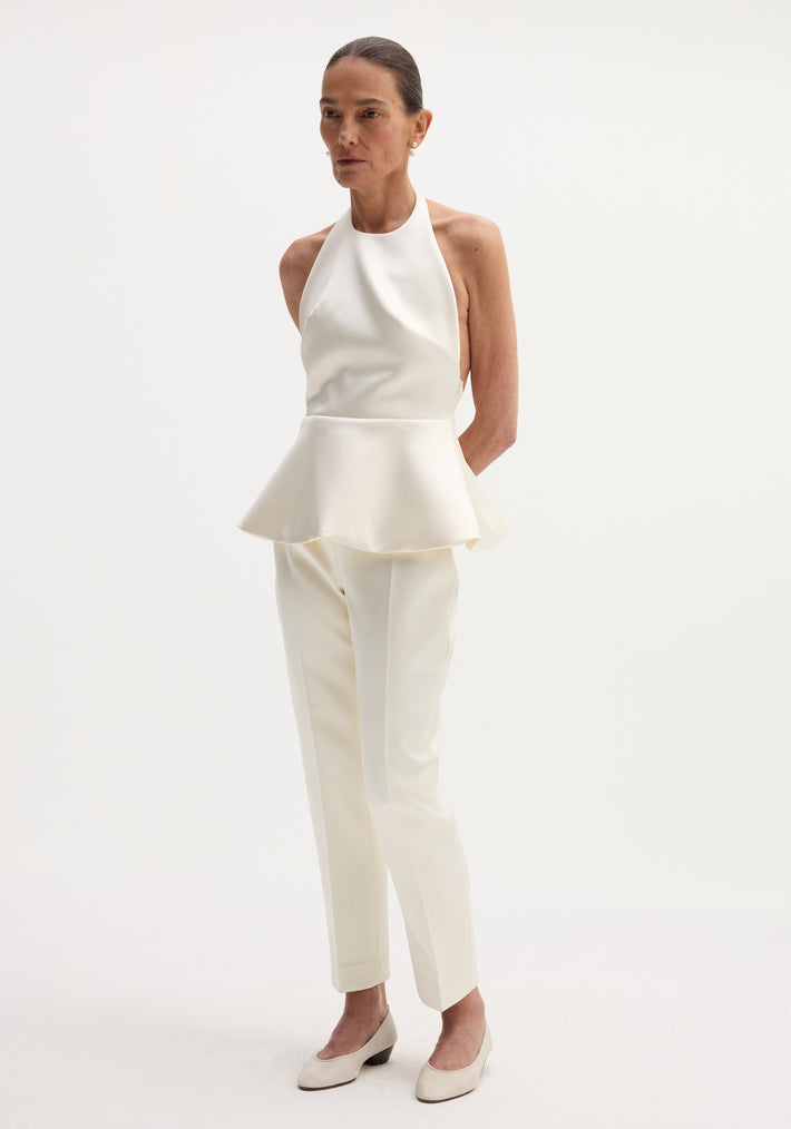 Model in Sculptural satin halter top with peplum, paired with straight-leg trousers and low-heeled pumps, showcasing minimalist fashion against a plain background.