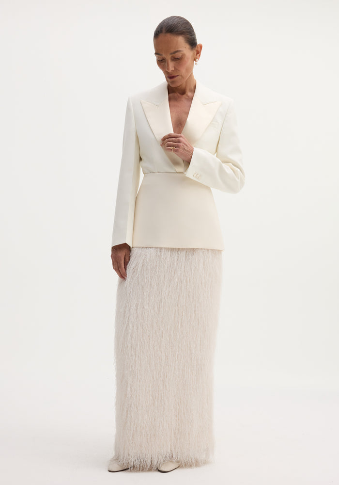 Woman wearing an ivory Fringe skirt with a structured wool upper and flowing fringe detail, touching her blazer lapel against a plain backdrop.