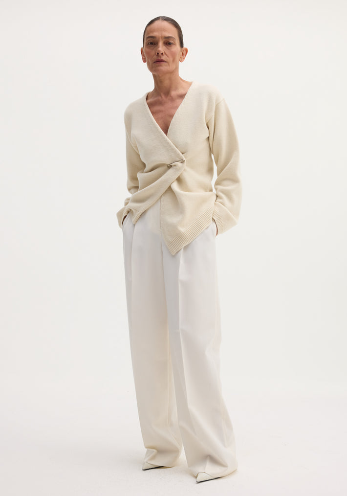 Off-shoulder wrap cardigan in cream, worn by a slender model, features a relaxed fit and metal safety-pin closure, paired with wide-leg trousers.