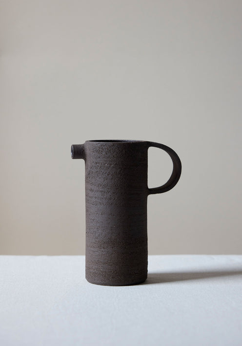 Ceramic Carafe | soil: A tall, dark, rough-textured ceramic carafe with a small spout and looped handle, showcasing a handcrafted, brutalist design by Alex Gabriels.