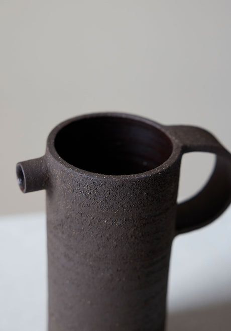 Ceramic Carafe | soil: A tall, dark, rough-textured ceramic carafe with a small spout and looped handle, showcasing a handcrafted, brutalist design by Alex Gabriels.