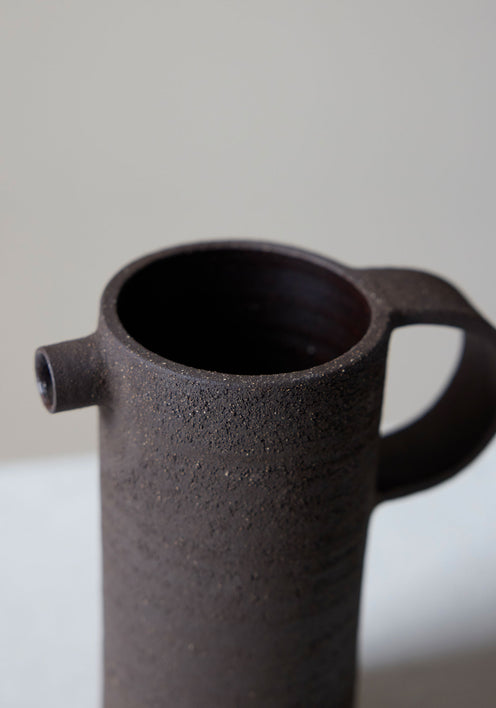 Ceramic Carafe | soil: A tall, dark, rough-textured ceramic carafe with a small spout and looped handle, showcasing a handcrafted, brutalist design by Alex Gabriels.