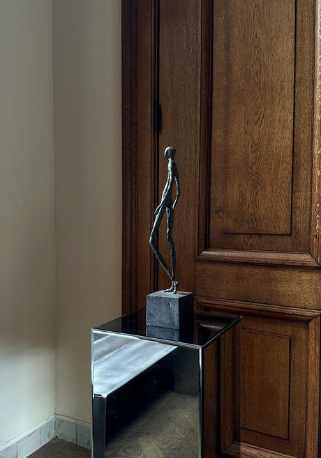 Bronze Sculpture | Quiet Confidence: An abstract bronze stick-figure sculpture on a black block, crafted by Dutch artist Veri Wisman, atop a mirrored cube pedestal.