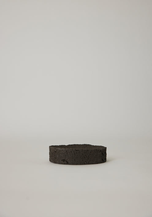 Round Tray made of textured lava stone, one on a pedestal, another holding wooden sticks, showcasing its handcrafted uniqueness on a white cloth.