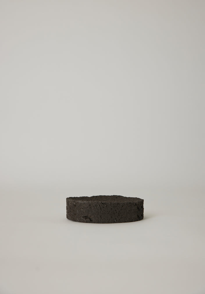 Round Tray made of textured lava stone, one on a pedestal, another holding wooden sticks, showcasing its handcrafted uniqueness on a white cloth.