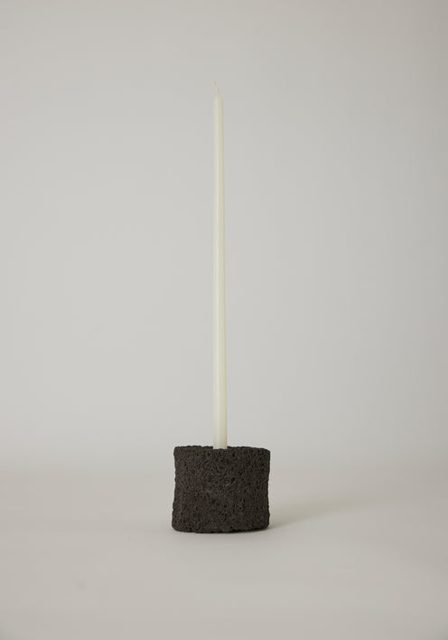 Round Candle Holder on minimal side table, crafted from lava stone, featuring a tall candle. Books and a stone accent the scene's serene, sparse design.