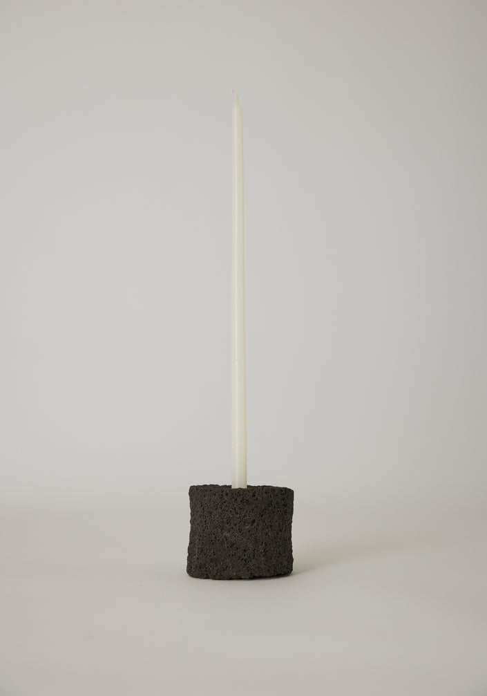 Round Candle Holder on minimal side table, crafted from lava stone, featuring a tall candle. Books and a stone accent the scene's serene, sparse design.