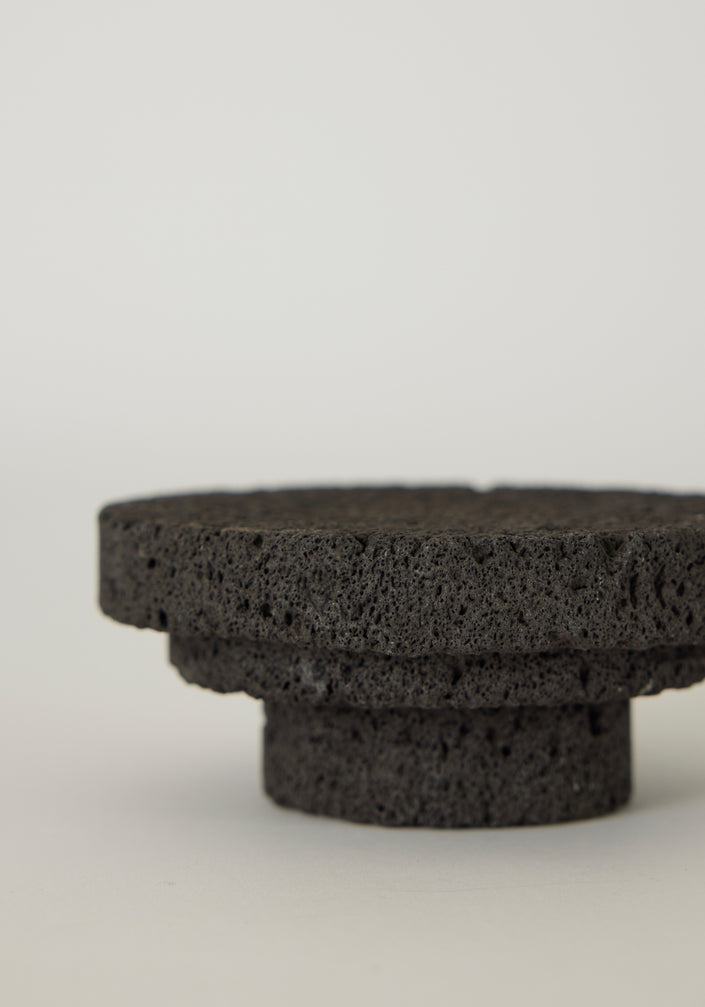 Elevated Sculptural Tray: A handcrafted, three-tiered circular pedestal made from dark lava stone by artist Louis Crombag, showcasing a unique, porous texture.