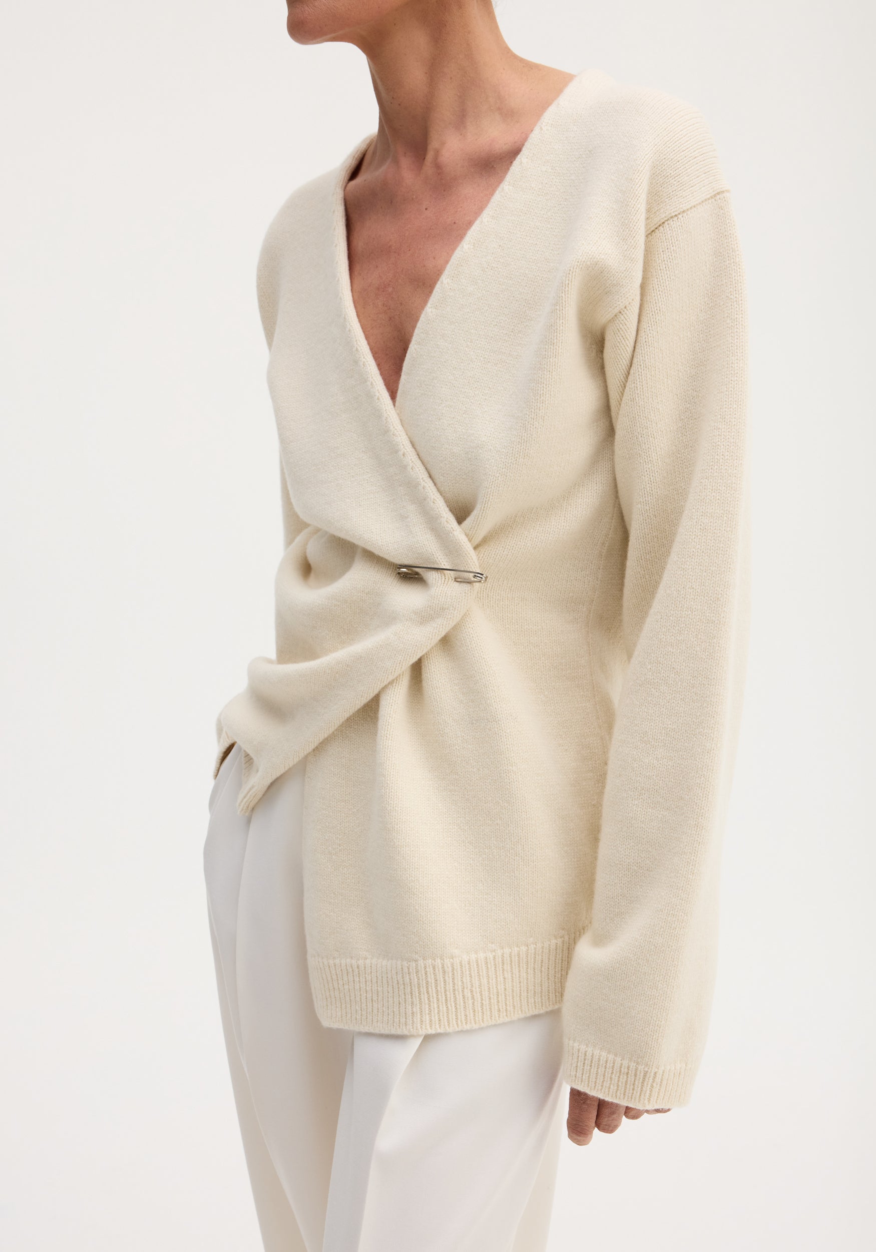 Woman wearing an off-shoulder wrap cardigan in cream, featuring a deep V-neck and metal pin closure, paired with high-waisted, wide-leg trousers.