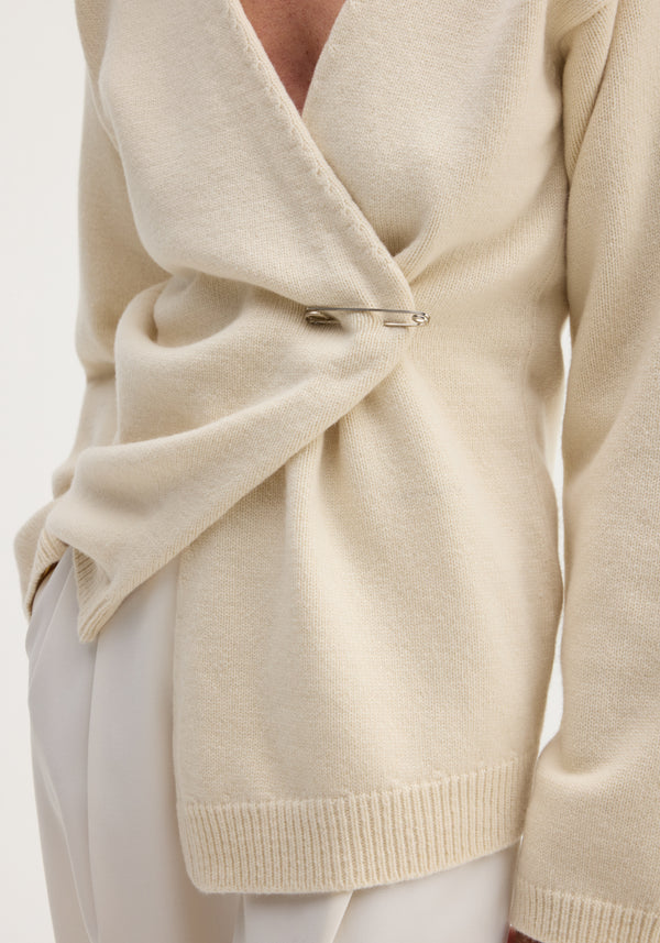 Off-shoulder wrap cardigan in cream, featuring a relaxed fit and secured with a large silver safety pin, paired with light-colored trousers.