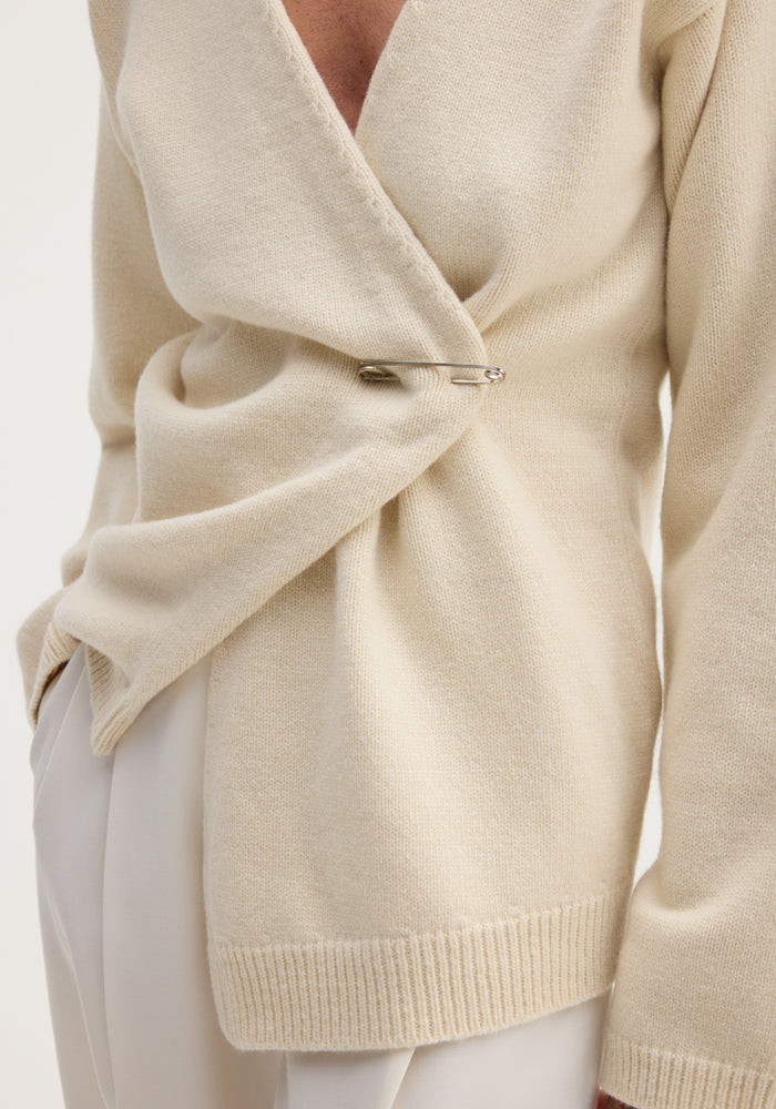 Off-shoulder wrap cardigan in cream, featuring a relaxed fit and secured with a large silver safety pin, paired with light-colored trousers.